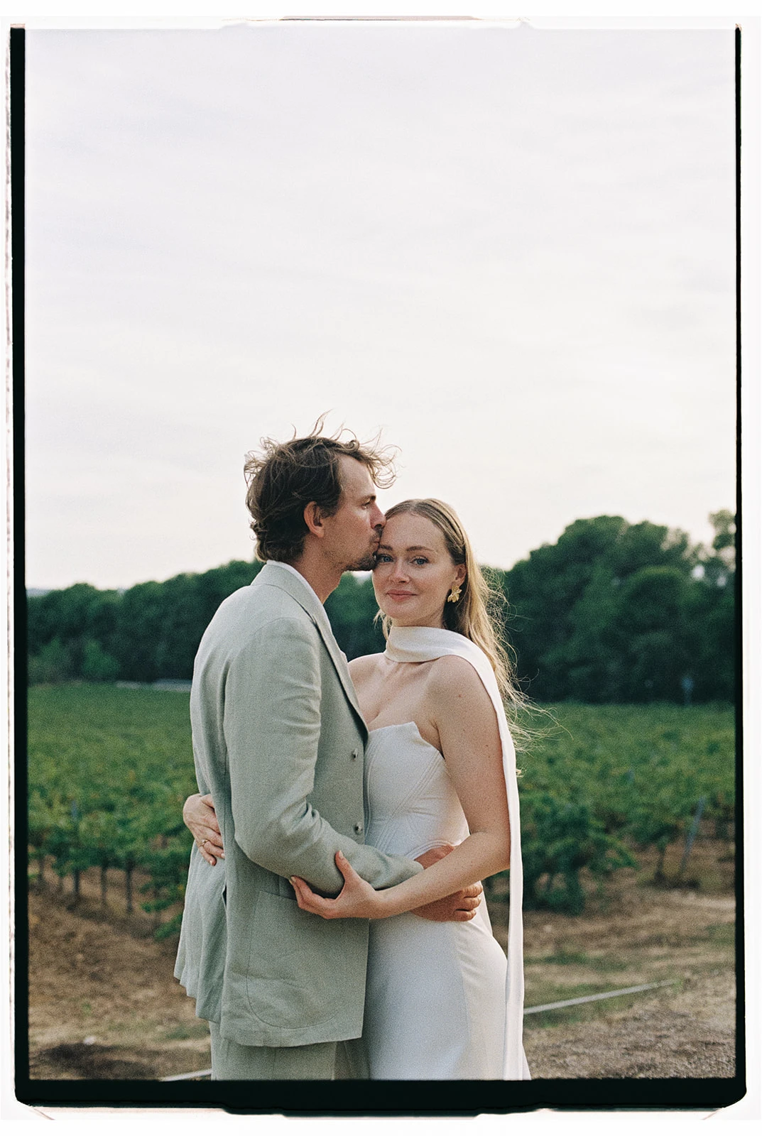Fine art film portrait of a groom in a sage green suit kissing a bride in a minimalist dress, set in a Mediterranean vineyard by Jessica North.