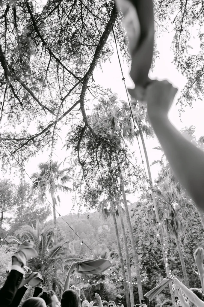 Black and white documentary photography of wedding guests waving napkins under palm trees and fairy lights in Mallorca. Festive celebration style