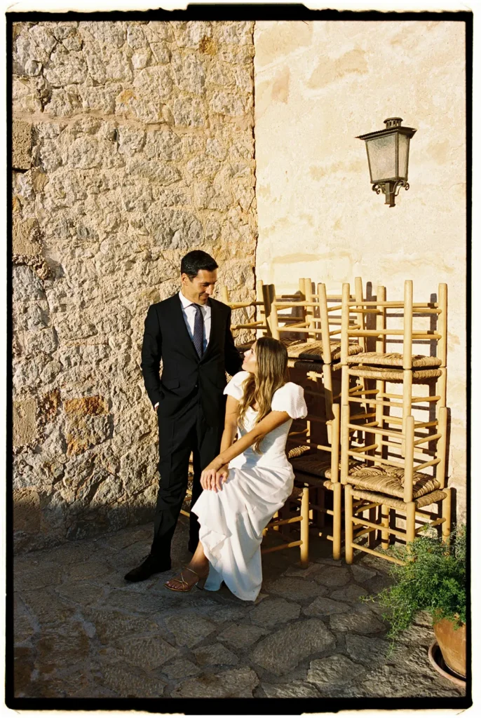 Fine art film photography of a bride and groom sitting by stacked chairs against a stone wall in a Mediterranean villa by Jessica North.